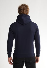 Marineblauwe hoodie van zacht katoen, met een grote capuchon, geribde manchetten en een aansluitende pasvorm. De achterkant toont een eenvoudig ontwerp en minimale branding.