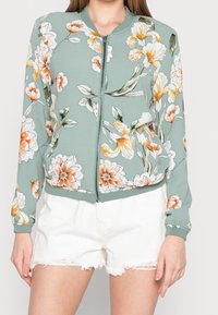 Chaqueta bomber floral verde con flores naranjas y blancas, cierre de cremallera en el frente, puños de canalé y dos bolsillos frontales. Llevada sobre pantalones cortos blancos.