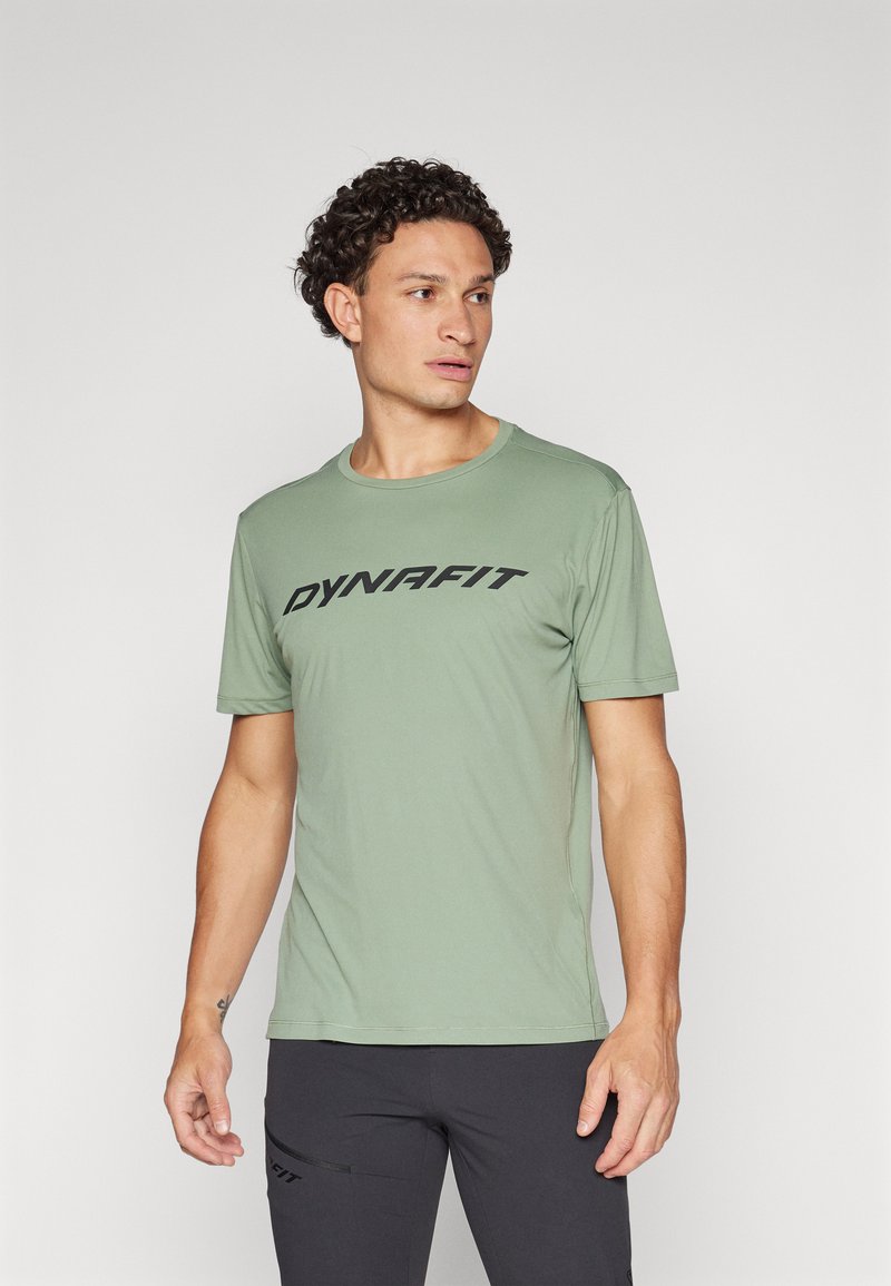 La camiseta atlética de color verde oliva presenta un escote redondo y mangas cortas, con un destacado logo negro "DYNAFIT" en el pecho.