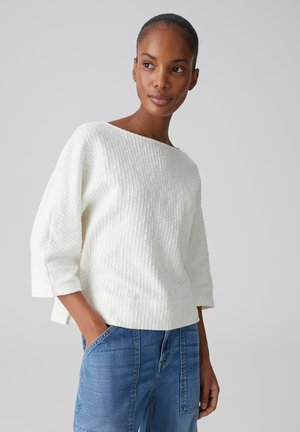 Frau trägt einen weißen strukturierten Pullover mit Bootsausschnitt und blaue Jeans, die Hände in den Taschen, blickt leicht zur Seite vor einfarbigem Hintergrund.