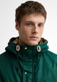 Νεαρός άνδρας με κοντά καστανά μαλλιά φορώντας πράσινο μπουφάν με κουκούλα και μπεζ κορδόνια, ουδέτερη έκφραση, απλό ανοιχτό φόντο.