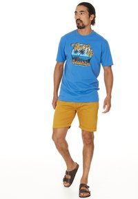 Mies kävelee päällään sininen "Surf's Up Beachlife" -t-paita, sinapinkeltaiset shortsit ja ruskeat solkisandaalit valkoista taustaa vasten.