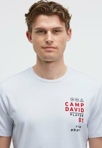 White short-sleeve t-shirt featuring red text "CAMP DAVID PLAYER 01" and graphic symbols. Smooth fabric with a classic crew neck design.