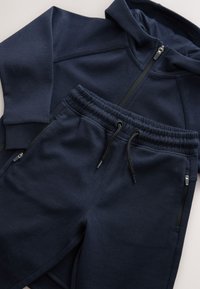 Felpa con cappuccio blu navy con zip frontale abbinata a pantaloni della tuta blu navy coordinati con coulisse e tasche con cerniera.