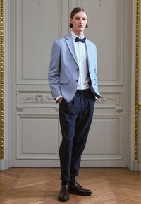 Gaiši pelēks uzvalka žakets ar teksturētu apdari, balta krekla, melna kaklasaites, tumšas brīvas bikses un brūni šņorējamie apavi. Koka grīdas fons.