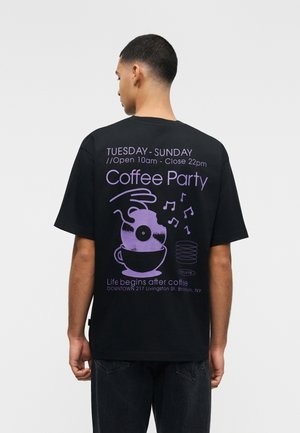 Mann trägt ein schwarzes T-Shirt mit lila Text und Grafiken, die für eine "Coffee Party" Veranstaltung von Dienstag bis Sonntag, 10 Uhr bis 22 Uhr, werben.