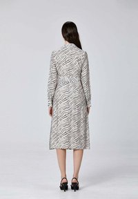 Robe longue beige avec un motif abstract noir. Elle possède des manches longues, une taille cintrée et une jupe fluide. Escarpins noirs à talons.