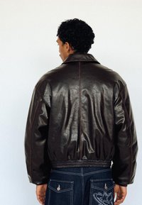 Man facing away wearing a dark brown leather bomber jacket and dark jeans with star embroidery on back pockets against a plain white background.