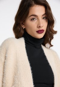Cremefarvet, fluffy cardigan med glitrende detaljer over en sort turtleneck. Krøllet hår og mørk læbestift komplementerer outfittet. Ren baggrund.