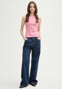 Różowy prążkowany top na ramiączkach zestawiony z szerokimi ciemnoniebieskimi dżinsami. Modelka stoi z dłońmi w kieszeniach. Gładki materiał i luźny krój.