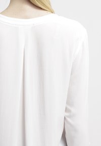 Hvid langærmet top lavet af letvægtsmateriale, med en afslappet pasform og et subtilt bagventdesign. Glat tekstur med minimalistisk syning.