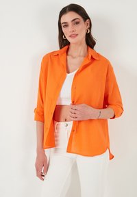 Světlá oranžová košile s límečkem, dlouhými rukávy vyhrnutými, nošená přes bílý crop top, spárovaná s bílými kalhotami. Hladká textura.