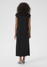 Robe maxi noire à manches courtes, avec une silhouette droite, un tissu lisse et une longueur jusqu'à la cheville, associée à des sandales à talons bleues.