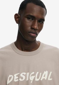 Beige tričko s krátkým rukávem, které má velké, strukturované bílé logo "DESIGUAL". Kulatý výstřih a hladký bavlněný materiál s volným střihem.
