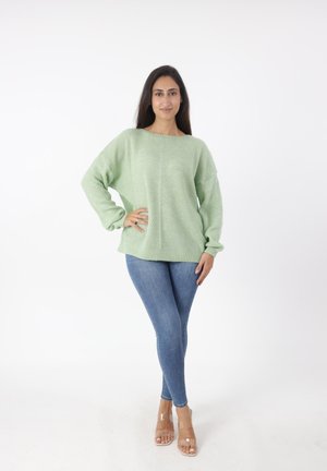 Maglione verde in maglia grossa con maniche lunghe e vestibilità comoda. Abbinato a jeans slim azzurri e sandali con tacco trasparente. Sfondo bianco semplice.