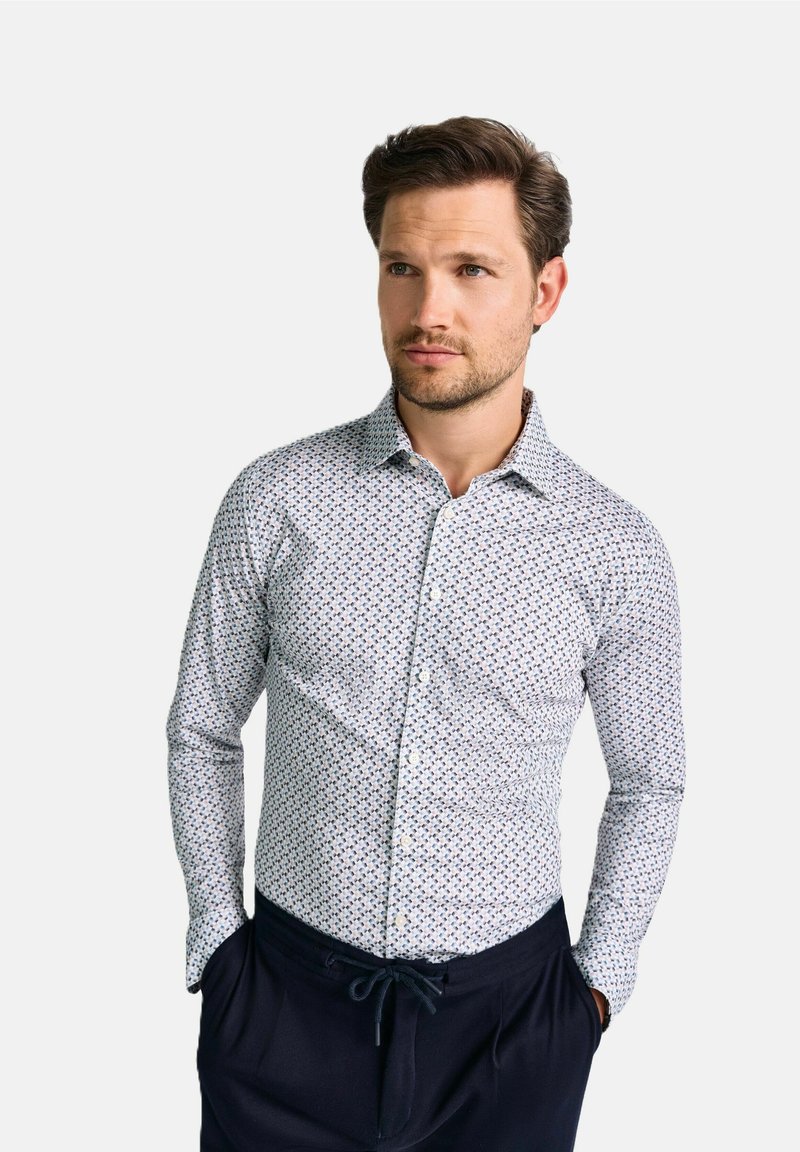 Weißes Hemd mit marineblauem und grünem Blumenmuster, Button-Down-Kragen, taillierter Schnitt, kombiniert mit dunkler Hose, Model steht mit Händen in den Taschen.