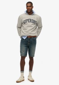 Felpa grigia con logo "SUPERDRY", colletto di camicia button-up blu, pantaloni corti in denim, sneakers beige e calze a righe crema. Design semplice.