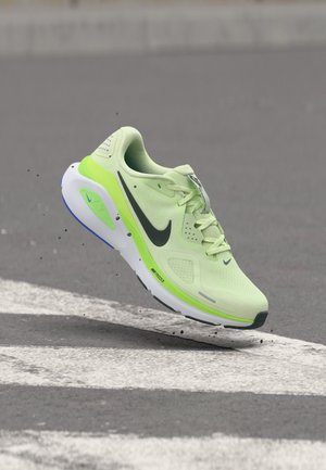 Jasnozielony but do biegania z czarnym logo Nike, białą i neonowozieloną podeszwą, lekko uniesiony nad szarą drogą z białymi oznaczeniami.