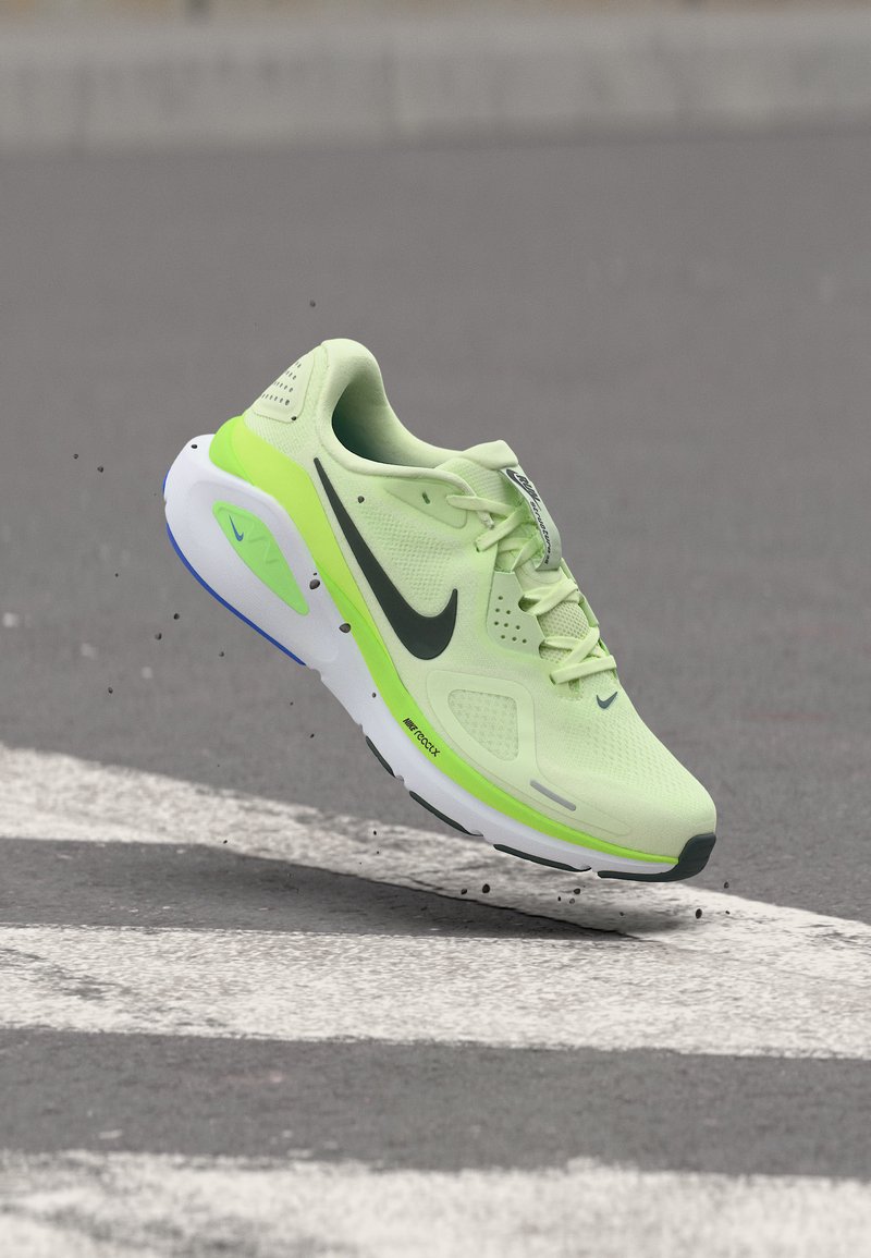 Ελαφρύ πράσινο αθλητικό παπούτσι με μαύρο λογότυπο Nike, λευκή και νέον πράσινη σόλα, ελαφρώς στον αέρα πάνω από έναν γκρι δρόμο με λευκές γραμμές.