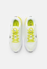 Bílé sportovní boty s žlutými akcenty, s texturovaným síťovaným vrškem, kulatými tkaničkami a černým logem Swoosh značky Nike na boku.