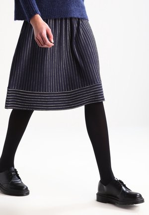 Person wearing a navy blue pinstripe skirt, black tights, black lace-up shoes, and a blue sweater, standing with legs apart on a white background.