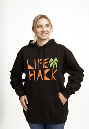 Donna sorridente, che indossa una felpa nero con cappuccio con la scritta "LIFE HACK" e una grafica di una palma verde, mani nelle tasche della felpa, su sfondo bianco uniforme.