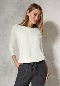 Weißes Langarmoberteil mit Rundhalsausschnitt und mittlerer Naht, kombiniert mit dunkelgrauen Jogginghosen mit Kordelzug. Weicher, leichter Stoff, lässiges Design.