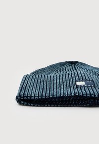 Sinine soonik-narmaskott, mille ees on Tommy Jeans logo kleebis, kui see on volditud, ja see on paigutatud lihtsa heleda tausta ette.