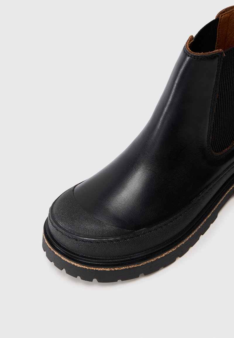 Bottine Chelsea en cuir noir avec un capuchon en caoutchouc texturé, panneau latéral élastique et semelle robuste sur un fond blanc.