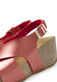 Red wedge sandal featuring a glossy leather upper with crisscross design and a cork-textured heel. Adjustable strap with silver buckle detail.