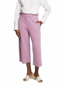 Femme portant un pantalon court lavande taille haute à jambes larges avec une taille plissée, associé à une chemise blanche à manches longues et des sandales beiges à talons blocs.