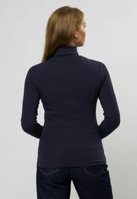 O femeie cu părul castaniu deschis, purtând un pulover cu guler înalt de culoare albastru marin, strâmt, și blugi închiși la culoare, stând cu fața întoarsă spre un fundal simplu.