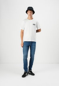Hvit t-skjorte med korte ermer og svart logo, blå slim-fit jeans, og svarte joggesko med hvite detaljer, kombinert med en svart bøttehatt.