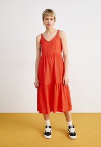 Robe maxi rouge sans manches avec un décolleté en V, jupe à volants et fines bretelles. Modèle portant des baskets noires et blanches avec des chaussettes blanches.