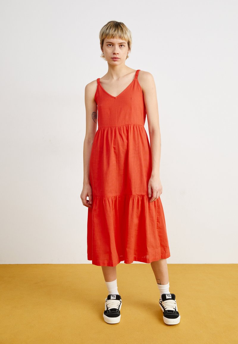 Robe maxi rouge sans manches avec un décolleté en V, jupe à volants et fines bretelles. Modèle portant des baskets noires et blanches avec des chaussettes blanches.