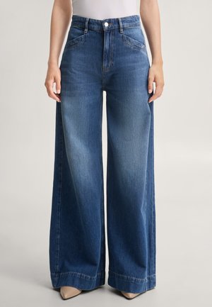 Persoon draagt blauwe high-waisted jeans met wijde pijpen en beige puntschoenen en staat tegen een egale lichte achtergrond.