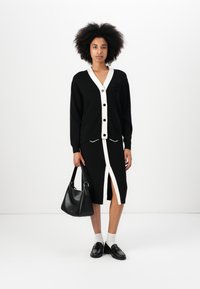 Musthave mustvalge triibuga ja nuppudega must kardigan, millega on sobitatud must seelik, millel on ees lõhik ja kreemi aktsendid. Tal on kaasas must käekott.