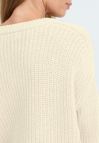 Lys kremfarget strikket genser med løs, teksturert veving, bred ribbestrikket halslinning og dropped shoulder-design, vist fra baksiden.