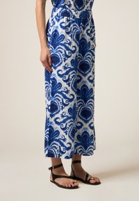 Blauwe en witte maxi jurk met een losvallende pasvorm en een elastische tailleband, gecombineerd met zwarte platte sandalen met gouden accenten.