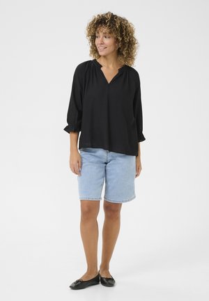 Blouse noire ample avec un col en V et des détails froncés au niveau du col, associée à un short en denim bleu clair. Des ballerines noires complètent la tenue.