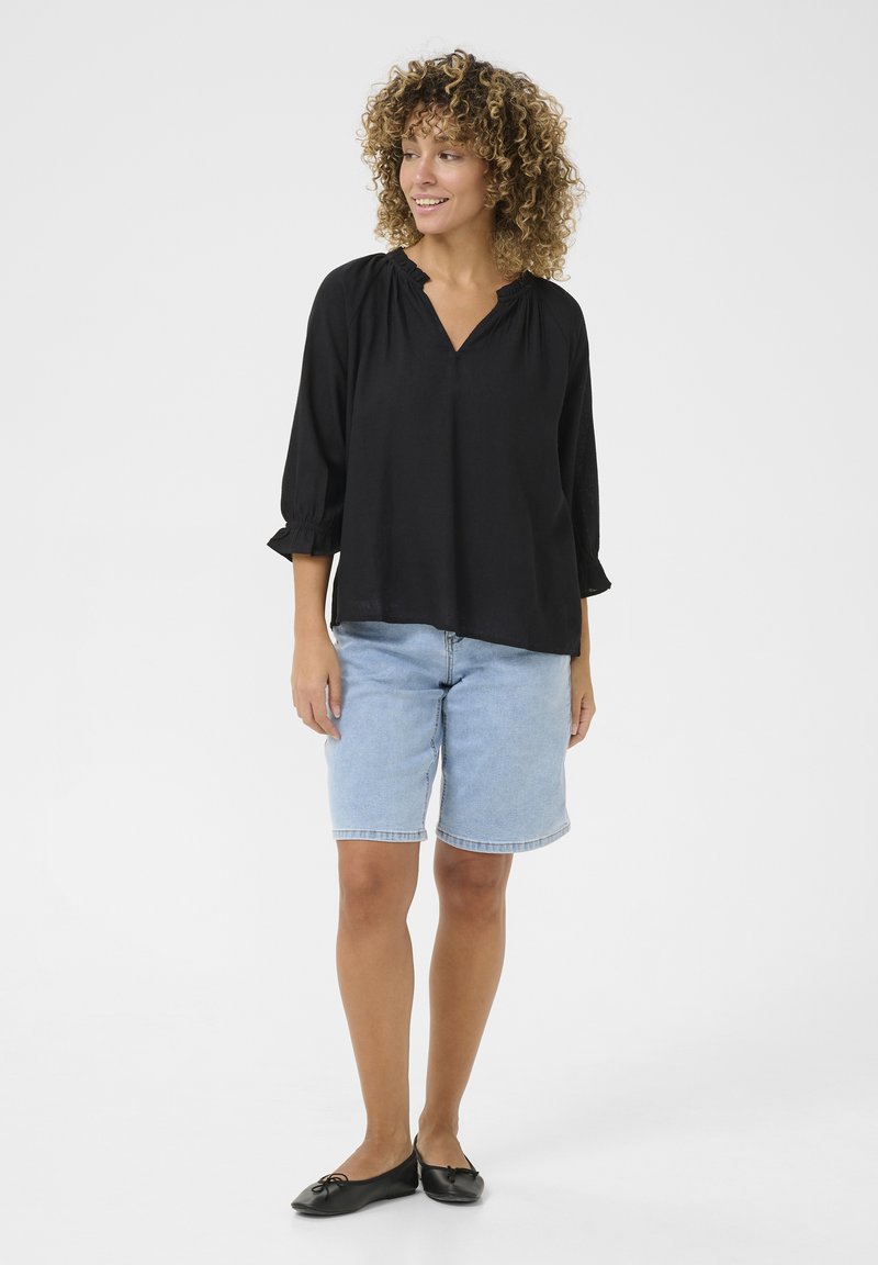 Schwarze, locker sitzende Bluse mit V-Ausschnitt und Raffung am Hals, kombiniert mit hellblauen Denim-Shorts. Schwarze Ballerinas runden das Outfit ab.