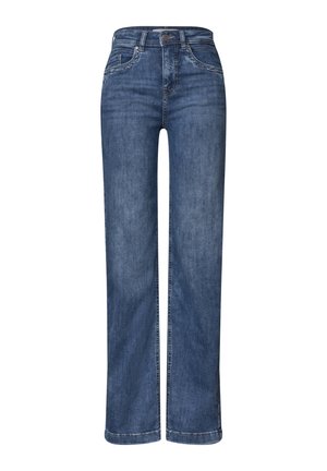Personne portant un jean en denim bleu, une chemise blanche et une veste bordeaux partiellement zippée, les mains détendues sur les côtés.