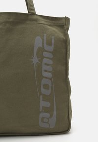 Sac fourre-tout en toile verte avec l'impression du logo "Atomic" en gris. Dispose de poignées robustes et d'une forme rectangulaire. Texture durable, adaptée à un usage quotidien.