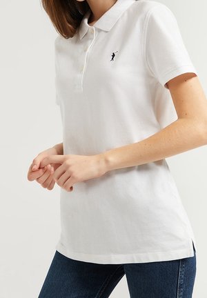 Kobieta nosząca białą koszulkę polo z krótkim rękawem i małym ciemnym haftowanym logo golfisty na lewej piersi, w połączeniu z ciemnymi dżinsami.