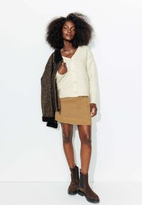 Cardigan en tricot crème avec encolure en V et fermeture à boutons, mini jupe en daim beige et bottines brunes. Le modèle tient une veste marron à imprimé léopard.