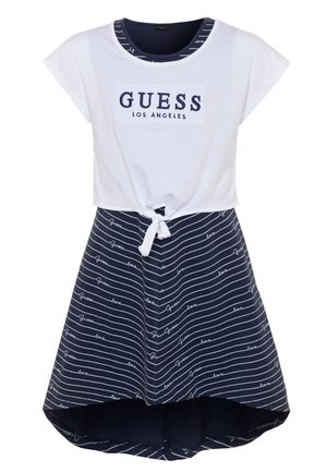 Biały top z krótkim rękawem z napisem "GUESS Los Angeles" związany w talii, w połączeniu z granatową spódnicą z białymi paskami oraz napisami "Guess" i "love".