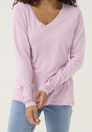 Kobieta w jasnoróżowej bluzce z długim rękawem i dekoltem w serek oraz niebieskich dżinsach, regulująca rękaw obiema rękami na wysokości talii.