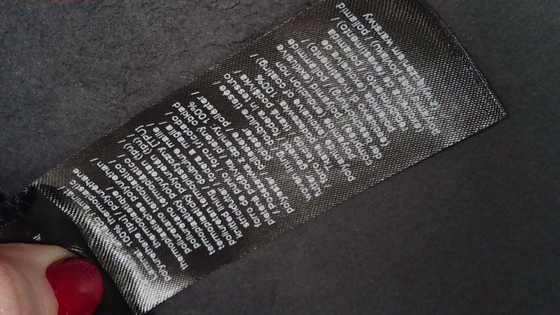 Czarna metka z instrukcjami prania, zawierająca białe napisy, szczegółowo opisująca skład materiału i zalecenia dotyczące pielęgnacji, o gładkiej, półprzezroczystej fakturze.