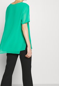 Blusa verde a maniche corte con una vestibilità ampia, scollatura rotonda e orlo a strati. Indossata con pantaloni neri aderenti.
