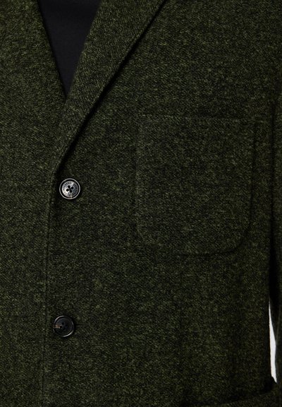 Mörkgrön ylleblazer med strukturerad tyg, en bröstficka och två svarta knappar fram för stängning.
